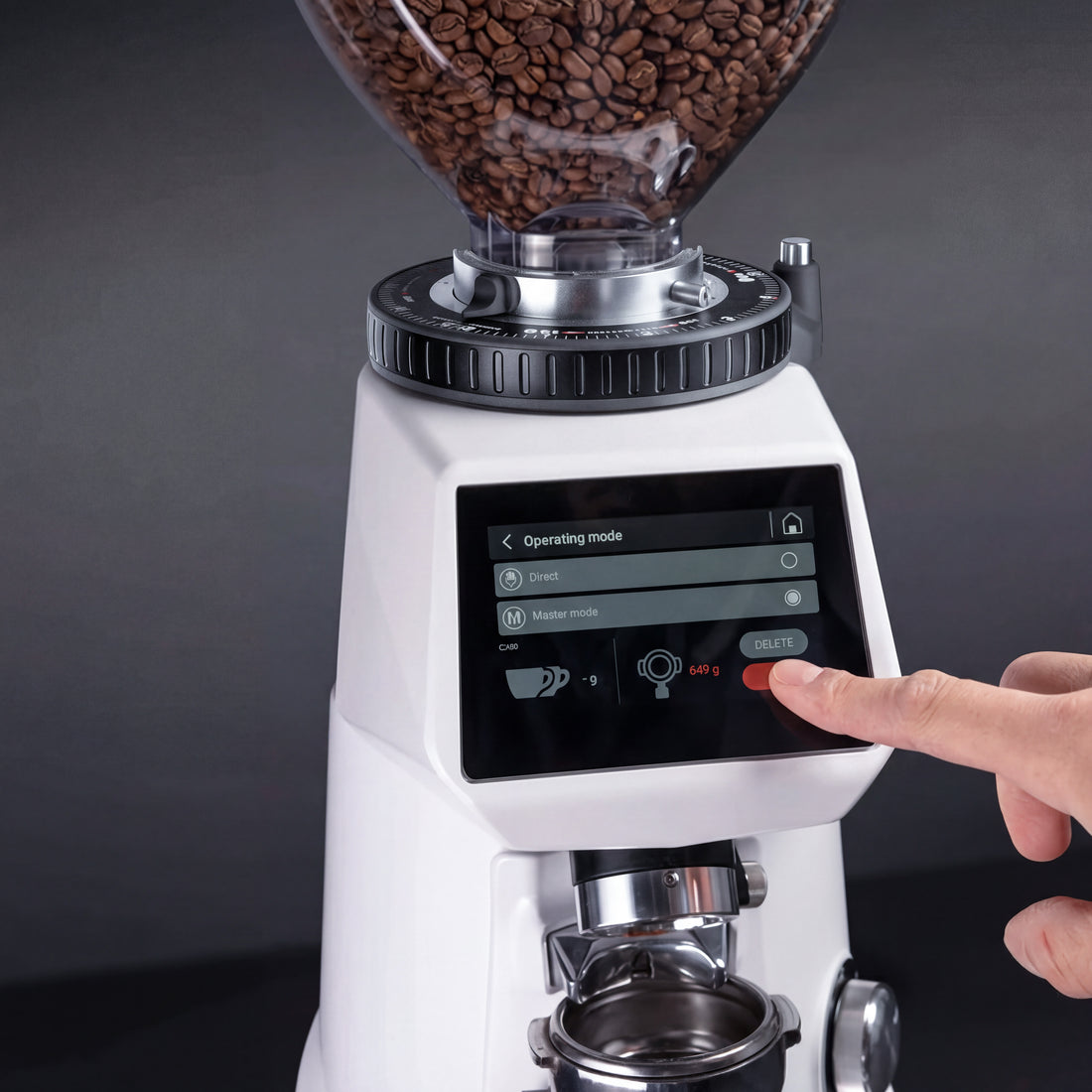 A close-up of a white electric coffee grinder filled with coffee beans, with a person's finger pressing the touch screen control panel. The grinder features a clear bean hopper, a black adjustment dial, and a portafilter holder at the bottom, set against a dark gray background.